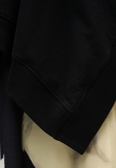 Črna jopa s rebrastim robom in teksturirano tkanino, prikazan je bližnji posnetek rokava in detajlov na manšeti.