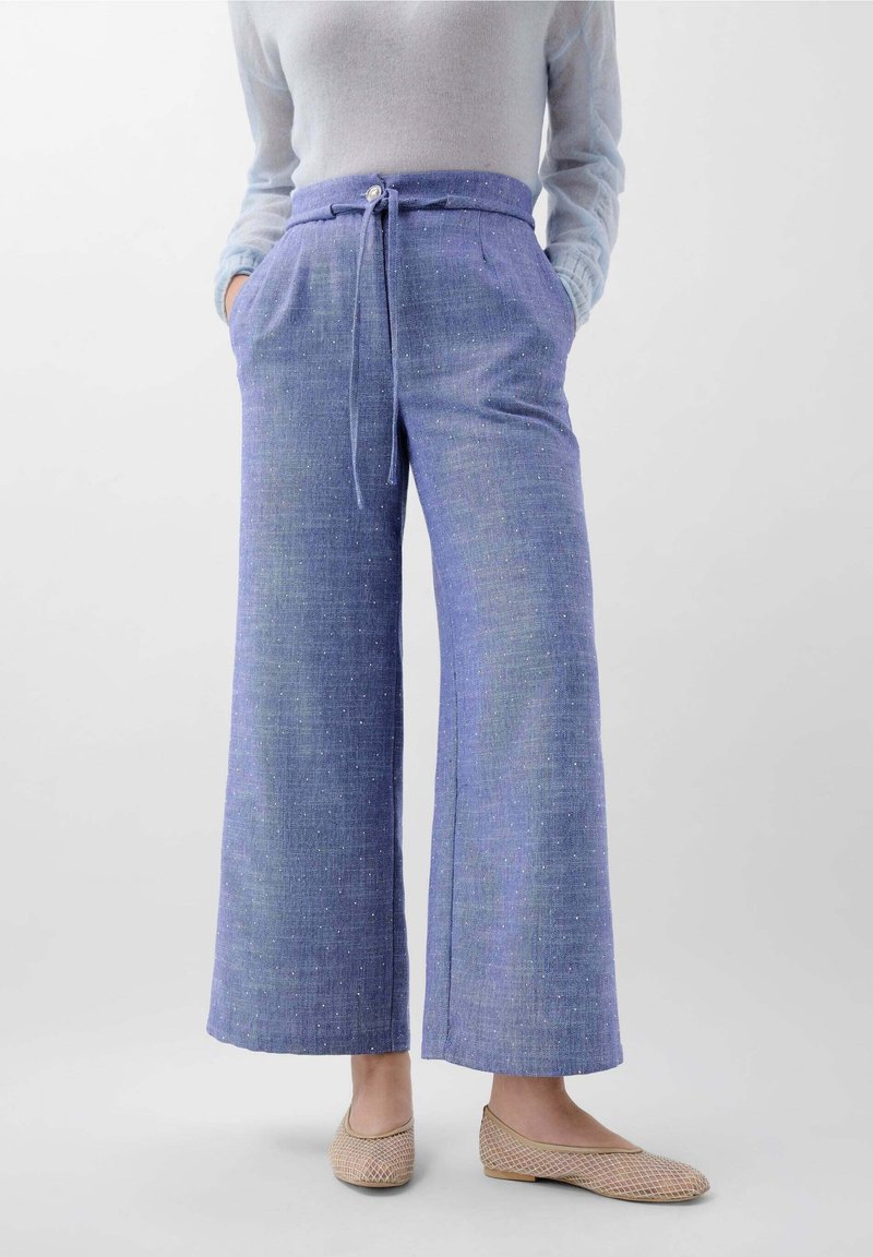 Femme portant un pantalon bleu taille haute à jambes larges avec une ceinture en tissu, combiné avec un haut blanc transparent à manches longues et des chaussures plates beiges.
