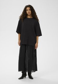 Schwarzes übergroßes T-Shirt mit weiten Ärmeln, kombiniert mit einem langen, transparenten schwarzen Rock und schwarzen Stiefeletten, vor einem schlichten Hintergrund.