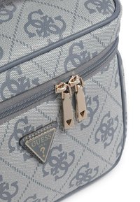 Primo piano di una borsa Guess grigia con motivo, dotata di due cursori in metallo argento e un'etichetta triangolare con il logo Guess.