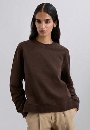 Sudadera marrón hecha de tela suave, con cuello redondo, puños de canalé y un sutil detalle de logo en el pecho. Combinada con pantalones beige.