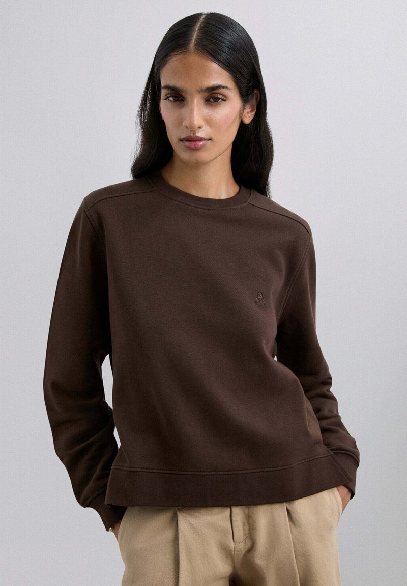 Sudadera marrón hecha de tela suave, con cuello redondo, puños de canalé y un sutil detalle de logo en el pecho. Combinada con pantalones beige.