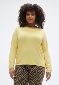 Rumeno pulover s dolgimi rokavi in okrolim izrezom, kombiniran s hlačami v rjavo-črnih tigrovih črtah, z udobnim krojem in mehko teksturo.