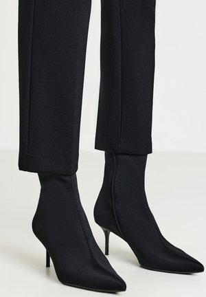 Stivaletti a punta nera con tacco a stiletto indossati con pantaloni neri a vita corta su uno sfondo chiaro.