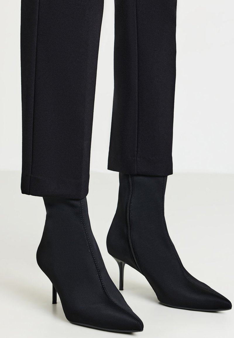 Stivaletti a punta nera con tacco a stiletto indossati con pantaloni neri a vita corta su uno sfondo chiaro.