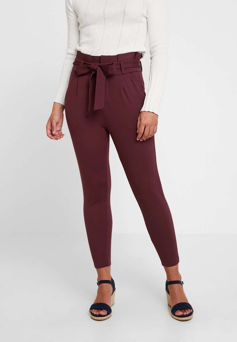 Pantalon bordeaux taille haute avec ceinture nouée, tissu lisse, coupe ajustée et poches latérales, associé à des sandales à talons bleu marine.