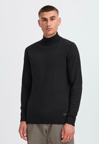 Pulover negru cu guler înalt, confecționat dintr-un material din tricot fin, având mâneci lungi, manșete cu nervuri și o patch logo subtil la tiv.