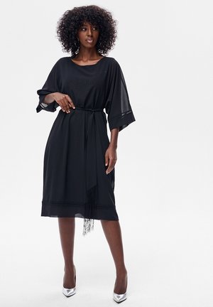 Kobieta o kręconych włosach, ubrana w czarną sukienkę midi z szerokimi rękawami i paskiem w talii, stojąca na srebrnych szpilkach na białym tle.