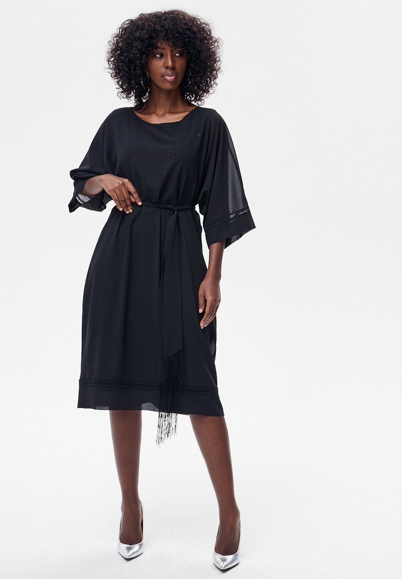 Kobieta o kręconych włosach, ubrana w czarną sukienkę midi z szerokimi rękawami i paskiem w talii, stojąca na srebrnych szpilkach na białym tle.