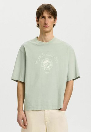 Joven con una camiseta suelta de color verde claro con texto en francés y pantalones beige, de pie con una mano en el bolsillo contra un fondo liso.