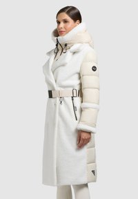 Bílý dvouřadý kabát s vycpávanými rukávy, vysokým límcem a páskem v pase. Texturovaná látka, hladké doplňky a logo na rukávu.