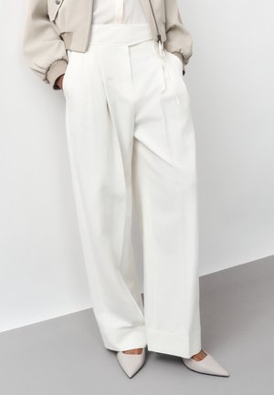 Γυναίκα που φοράει λευκό παντελόνι ψηλόμεσο με φαρδιά μπαλαντζέ πόδια, ένα μπεζ κοντό σακάκι και γκρι ανοιχτά μυτερά τακούνια, στέκεται μπροστά σε έναν λευκό τοίχο.