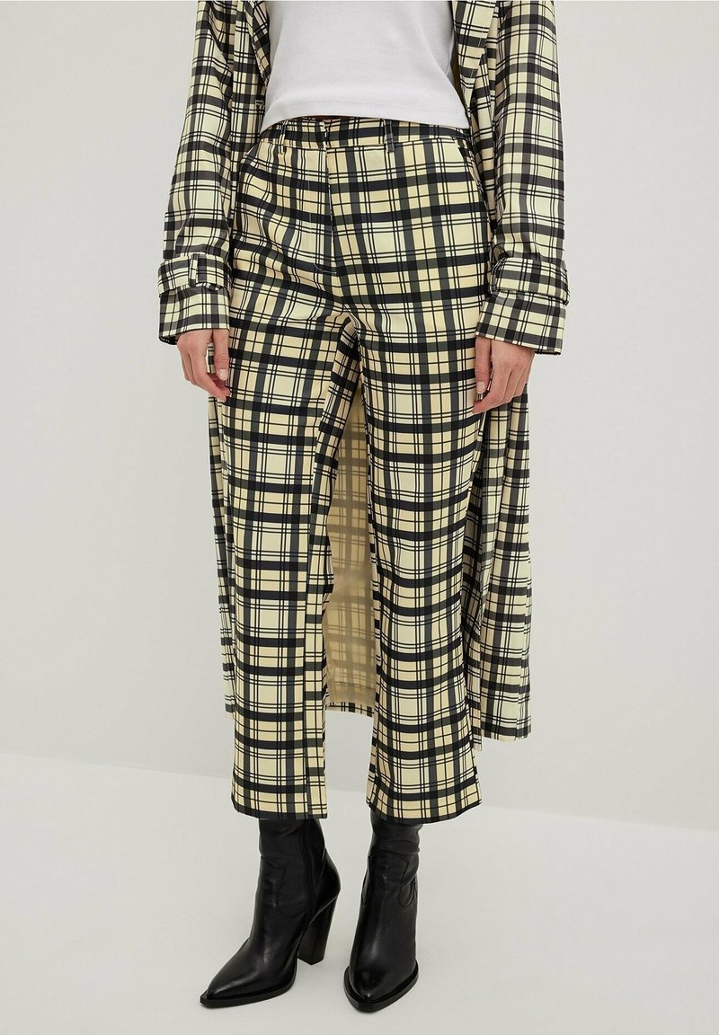 Femme portant un pantalon large à carreaux beige et noir, un manteau long assorti, un haut blanc et des bottines à talons noires, debout sur un sol blanc.