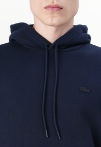 Tamsiai mėlynas džemperis su gobtuvu, pagamintas iš gumuoto audinio, turintis kengūros kišenę ir reguliuojamas raišteles su sidabro spalvos galiukais. Juodas Lacoste logotipas.