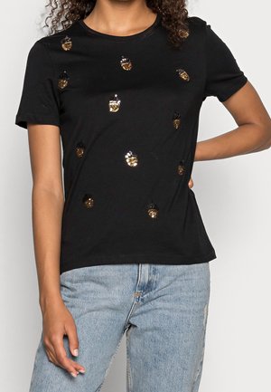 Czarny t-shirt z krótkim rękawem z wzorami żołędzi z złotych i czarnych cekinów, noszony z jasnoniebieskimi dżinsami, dłoń modelki na biodrze i widoczne kręcone włosy.