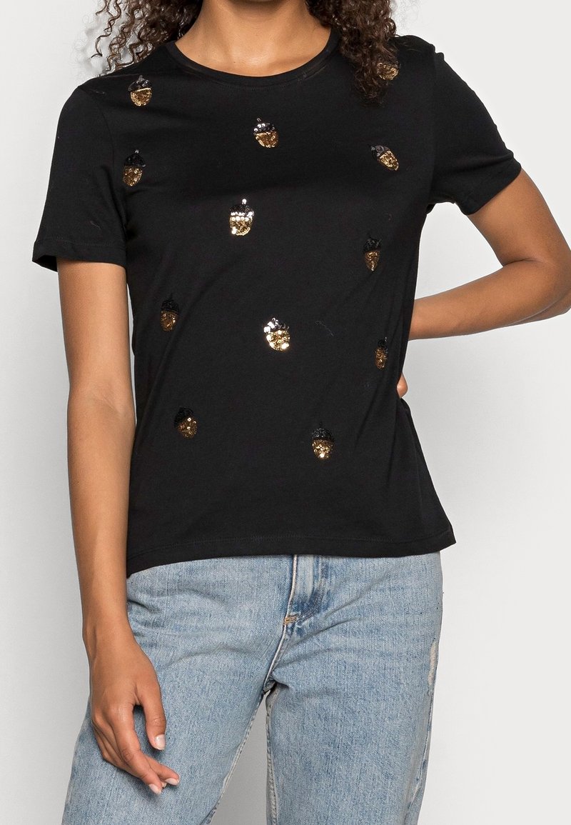 T-shirt noir à manches courtes avec motifs de glands en sequins dorés et noirs, porté avec un jean bleu clair, main du mannequin sur la hanche et cheveux bouclés visibles.