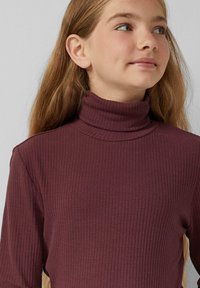Junges Mädchen mit langen Haaren, das einen gerippten kastanienbraunen Rollkragenpullover trägt und leicht nach oben und zur Seite blickt, vor einem grauen Hintergrund.