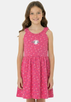 Fille souriante, portant une robe rose sans manches avec de petits motifs géométriques blancs et bleus et un dessin de cœur sur la poitrine.