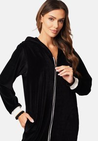 Černý sametový overall s kapucí, se zipovým uzávěrem a bílými pruhy na manžetách, hladký povrch a boční kapsy.