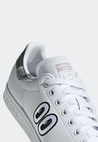 Baskets en cuir blanc avec un motif brodé noir et blanc sur le côté. Présente un accent pastel sur le talon et des lacets blancs.