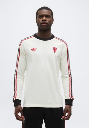 Hombre vistiendo camisa blanca de manga larga con rayas rojas y negras en las mangas y logotipos rojos en el pecho, posando frente a un fondo liso.