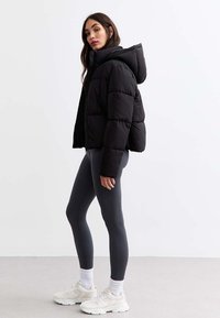 Czarna kurtka puchowa z kapturem, o pikowanym wzorze. Noszona na szare legginsy i białe sneakersy z teksturowanymi akcentami.