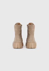 Paire de bottines en daim beige avec des semelles en caoutchouc épaisses et texturées, montrées de derrière sur un fond blanc uni.