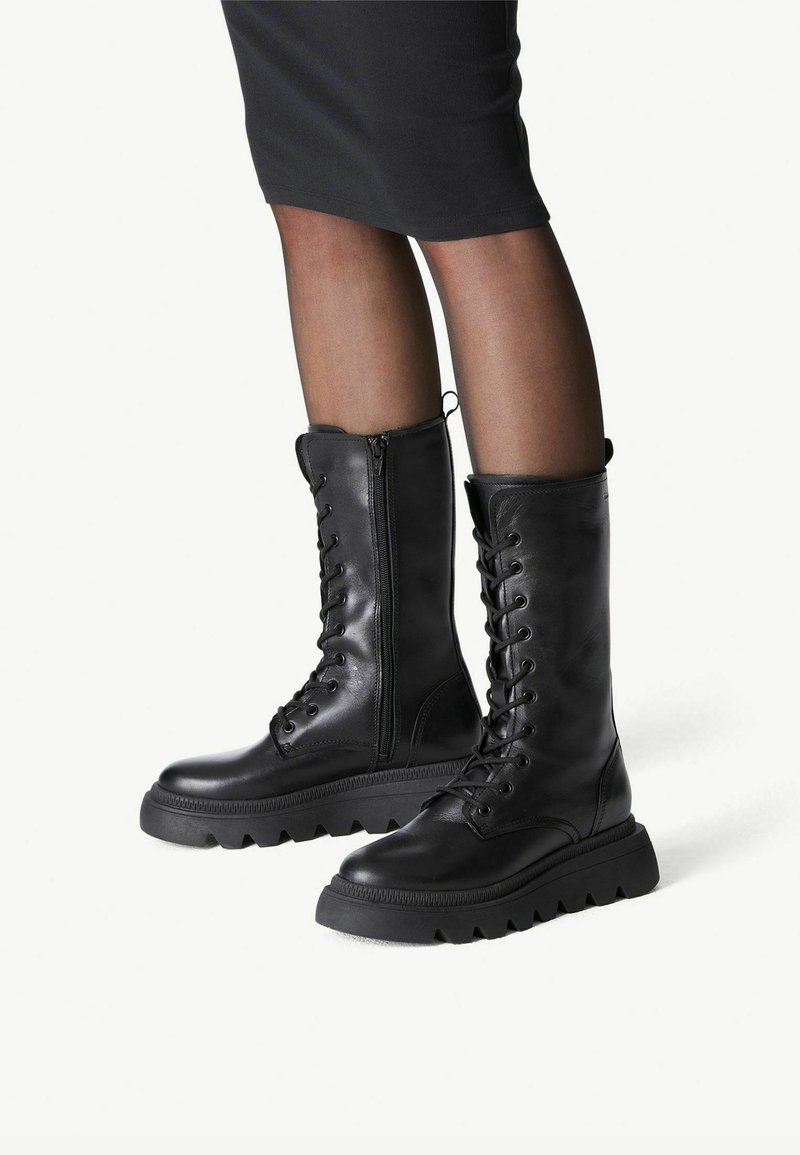 Bottes de combat en cuir noir montant jusqu’au milieu du mollet, avec des semelles épaisses et dentelées, portées avec des collants noirs transparents et une jupe sombre.