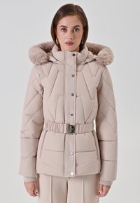 Vrouw in een beige gewatteerde pufferjack met een met bont afgewerkte capuchon en een ceintuur op de taille, staand tegen een effen lichte achtergrond.