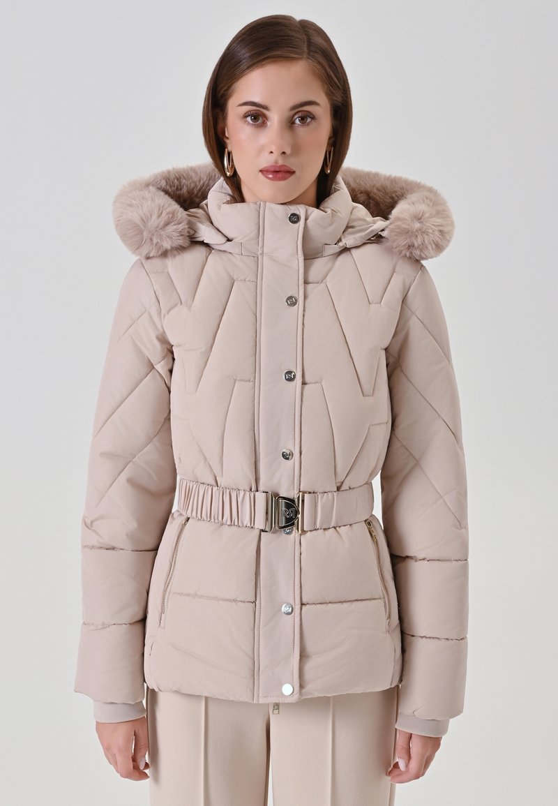 Vrouw in een beige gewatteerde pufferjack met een met bont afgewerkte capuchon en een ceintuur op de taille, staand tegen een effen lichte achtergrond.