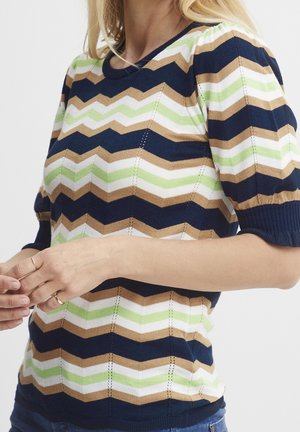 Mujer con un top de punto de manga corta con rayas de zigzag en azul marino, beige, blanco y verde claro, manos apoyadas al frente, recortada a la altura del cuello y las caderas.