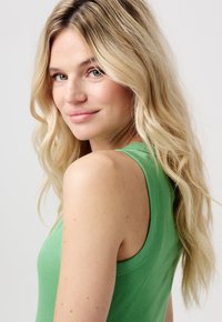 Blonde vrouw met lang golvend haar in een mouwloze groene top, zacht glimlachend, kijkt over haar schouder tegen een eenvoudige lichte achtergrond.