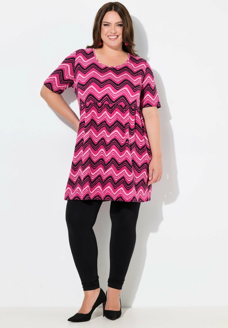 Tunika se zigzagovým vzorem v odstínech růžové a černé, krátké rukávy, volný střih, doplněná černými legínami a černými vysokými podpatky.
