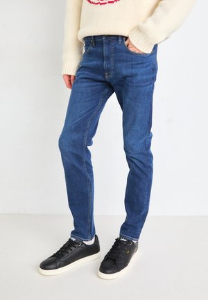 Blue denim jeans with a slim fit, featuring five pockets and a classic design. Worn with black sneakers and a cream knit sweater.