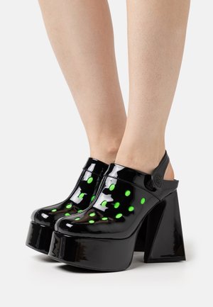 Sabots à plateforme en cuir verni noir avec un talon bloc, ornés de pois verts, à dos ouvert, avec une étiquette logo sur le côté.