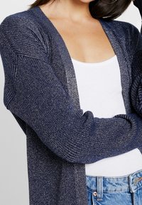 Námořnický cardigan s texturovaným, žebrovaným vzorem a metalickými nitěmi. Design s otevřeným předním dílem, kombinovaný s bílým topem a světle modrými džíny.