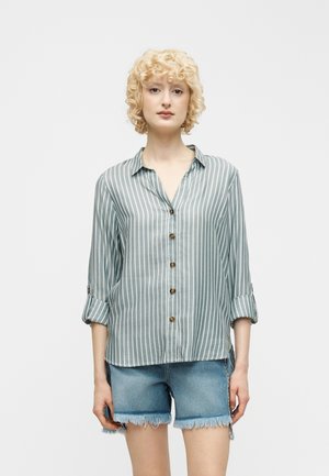 Femme blonde portant une chemise boutonnée à rayures verticales bleu clair et blanc, manches retroussées, et un short en jean effiloché.