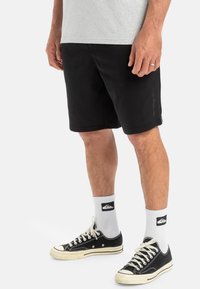 Pantalones cortos negros de material suave, con un ajuste relajado, bolsillos laterales y un logo sutil en la pierna. Combinados con zapatillas negras y calcetines blancos.