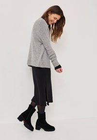 Suéter gris de punto con mangas largas, combinado con una falda negra hasta la rodilla que presenta una abertura lateral. Botines negros de ante con un tacón grueso.