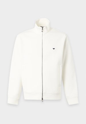 Homme portant une veste blanche zippée, un pantalon noir et des baskets blanches, debout devant un fond blanc uni avec une main dans la poche.