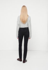 Mujer con un suéter gris y jeans ajustados negros, con cabello largo y zapatos marrón, vista de espaldas sobre un fondo blanco.