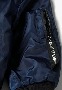 Mörkblå bomberjacka med en glänsande, slät textur. Har en ficka med dragkedja och ett svart tygband märkt "TAKE IT EASY."