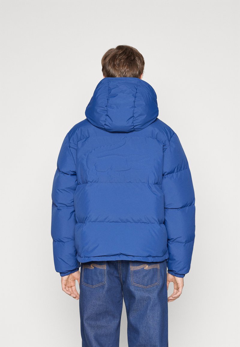 Hombre con una chaqueta acolchada azul que tiene un sutil logo de cocodrilo en la parte posterior, combinada con jeans azul oscuro, de espaldas a la cámara.
