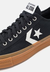 Schwarzer Segeltuch-Sneaker mit weißem Sternlogo, schwarzen Schnürsenkeln, silbernen Ösen und brauner Gummisohle. Klassisches Low-Top-Design.