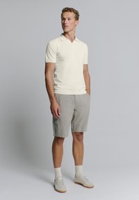 Lichtbeige polo shirt met een V-hals en korte mouwen, gecombineerd met grijze linnen shorts. Het model draagt witte sokken en lichte sneakers.