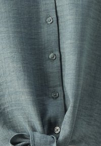 Nahaufnahme eines blau-grauen Stoffhemdes mit sichtbaren Knöpfen und einem gebundenen Knoten am Saum.