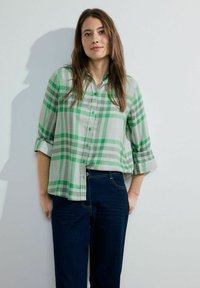 Camicia a quadri verde e grigia in cotone leggero, con un'apertura a bottoni e maniche arrotolate, abbinata a jeans blu scuro.