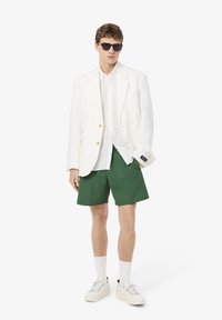Blazer en lin blanc sur une chemise claire, associé à un short vert. Les accessoires comprennent des lunettes de soleil noires et des baskets blanches avec des chaussettes hautes.