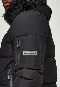 Czarna kurtka puchowa z kapturem wyłożonym futerkiem. Posiada kieszenie na zamek oraz naszywkę z logo na rękawie. Materiał o fakturze z pikowanym wzorem.