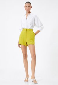 Camisa blanca de botones, pantalones cortos amarillos, sandalias planas. Los pantalones cortos tienen una cintura alta y una textura suave. Diseño sencillo, ajuste casual.
